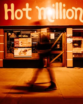 Blurred pedestrian walking past a brightly lit Hot Million storefront at night.