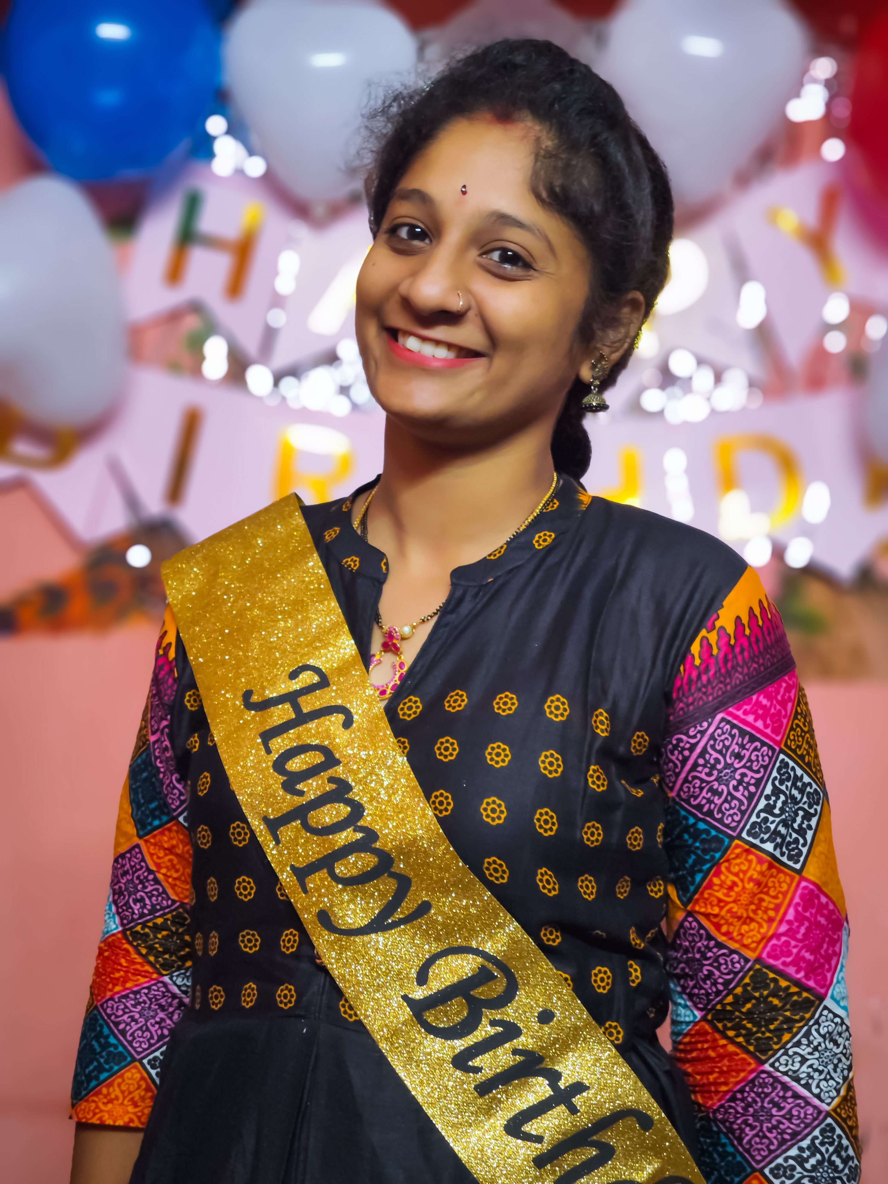 Smiling woman at a birthday celebration wearing a colorful dress and a Happy Birthday sash.