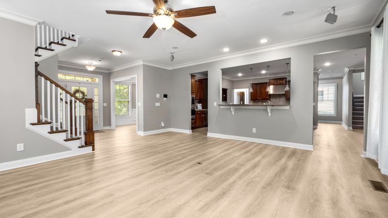 A modern ceiling fan spinning in a bright, sunlit living room with the wall thermostat visible in the background