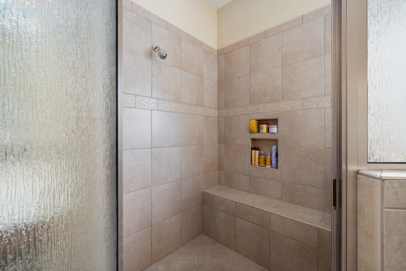 Enclosed beige tiled shower with a decorative horizontal border, recessed storage niche, built-in bench and textured glass door.
