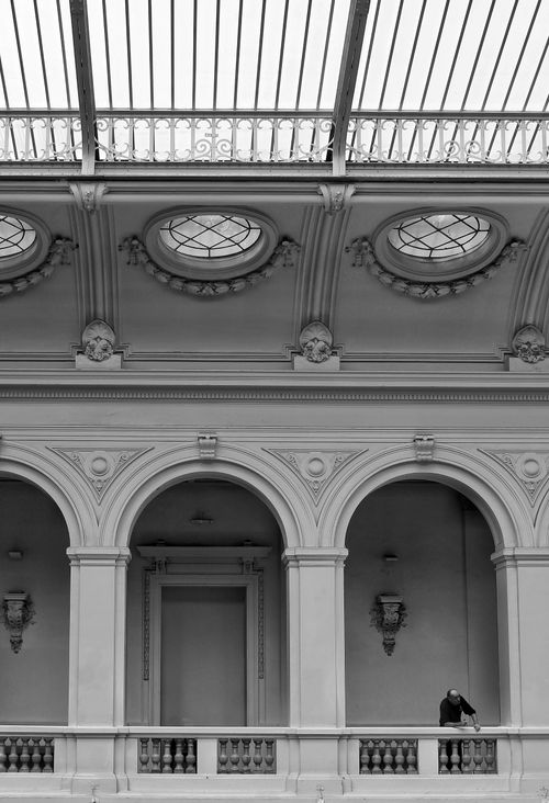 Free Classic black and white shot of an elegant interior balcony featuring ornate arches and a man standing alone. Stock Photo