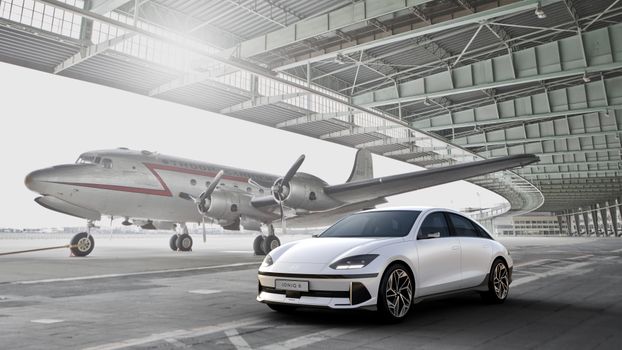Sleek white electric car parked near a vintage aircraft in an open hangar.