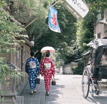 Two women wearing kimonos walk down a traditional street in Kyoto under a parasol, evoking cultural elegance.