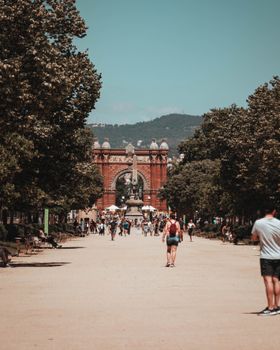 People walking on a sunny day near the Arc de Triomf in Barcelona, Spain. Trees line the path.