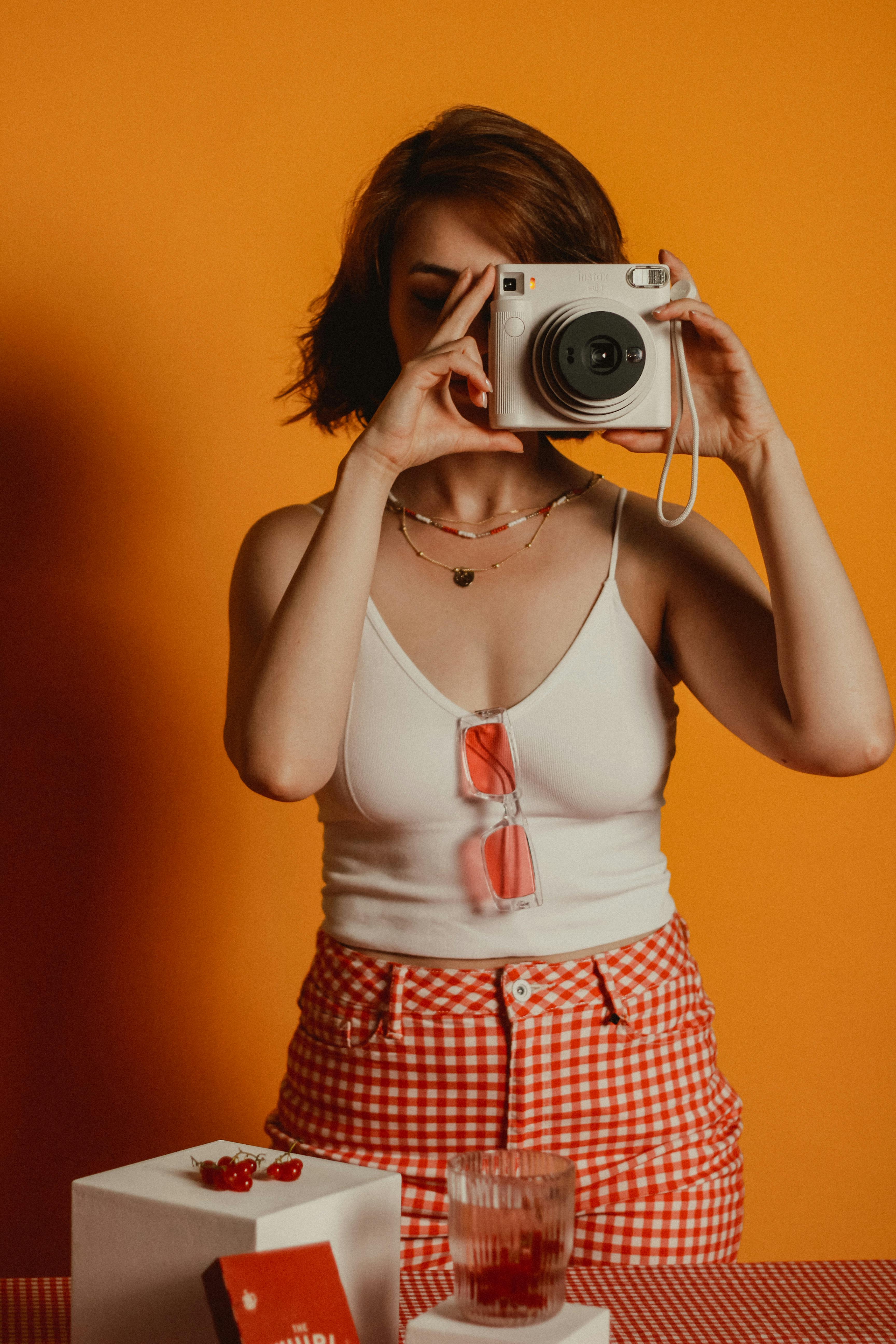 Woman holding instant camera with orange background in a studio setting.