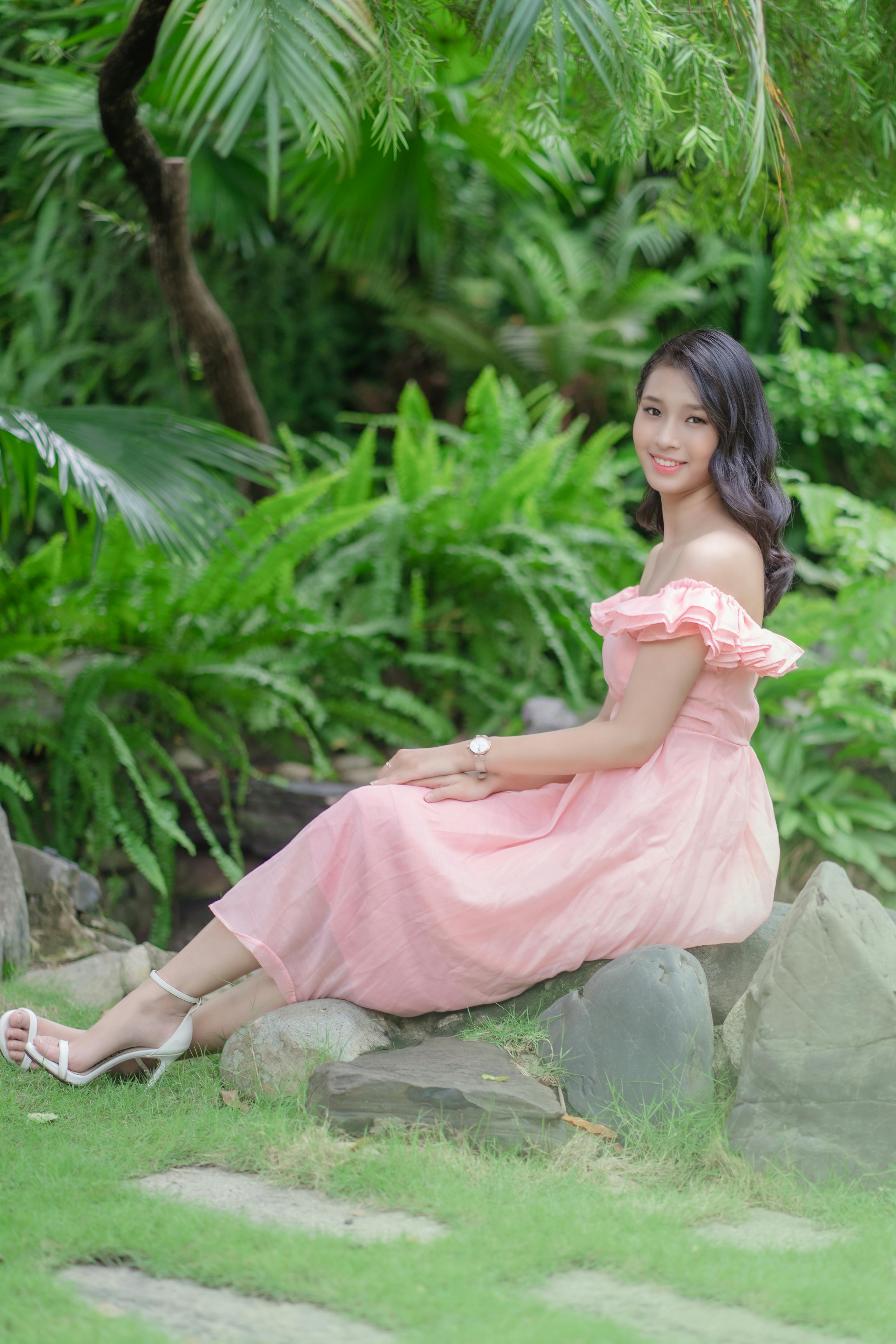 A smiling woman in a beautiful pink dress sits serenely among vibrant green foliage, exuding grace.