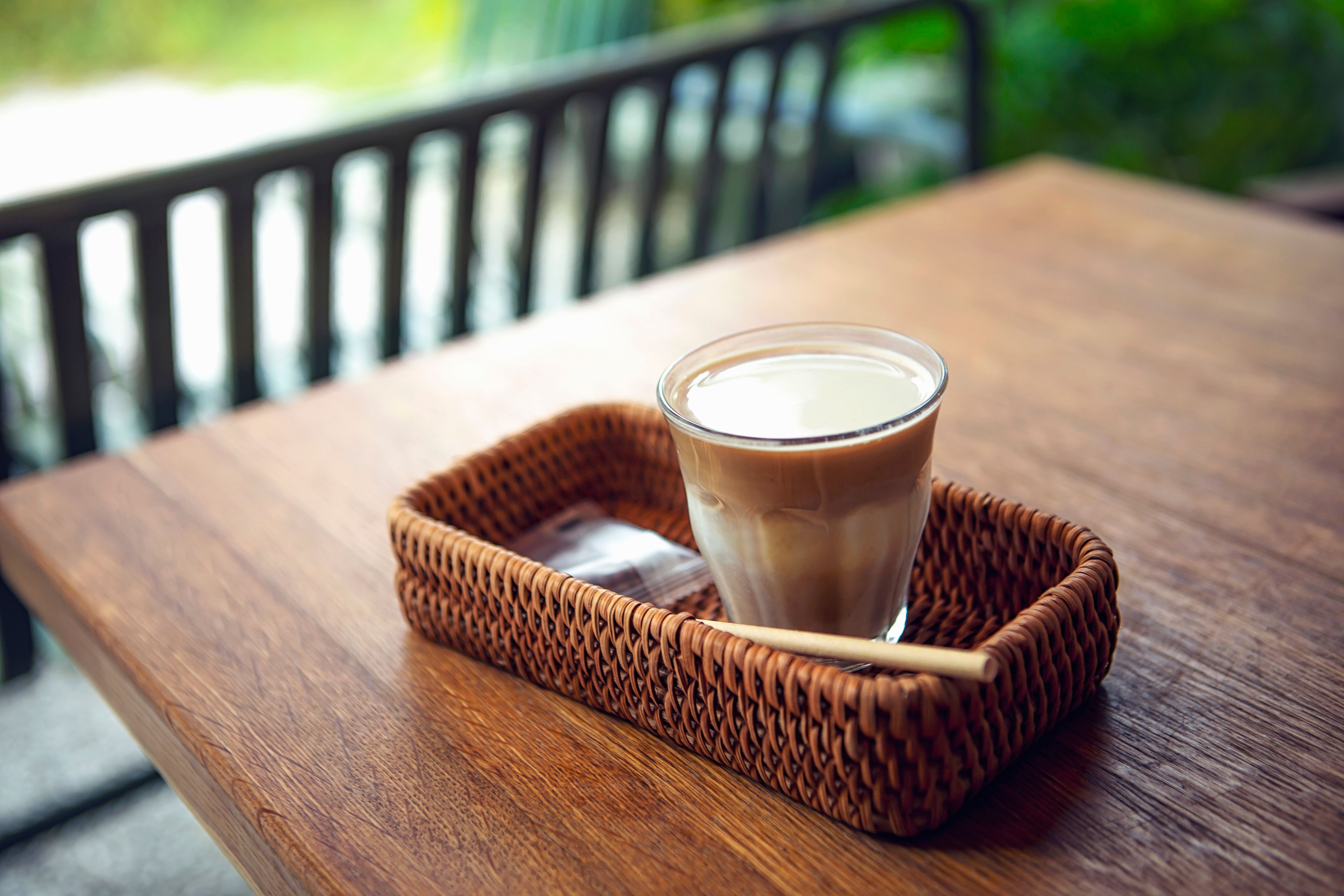 A cozy latte served in a glass within a woven basket on a wooden table outdoors.