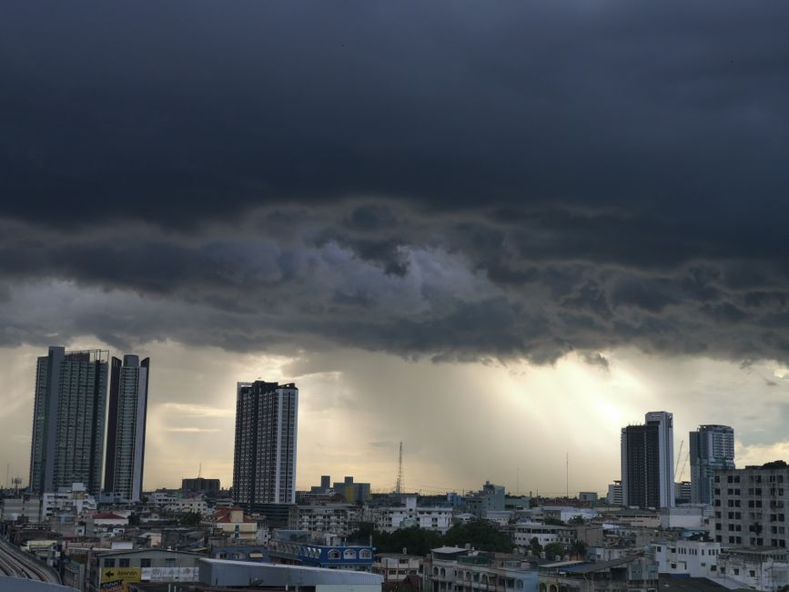 A dramatic cityscape view with tall buildings under a stormy sky with rain and sunlight peeking through.