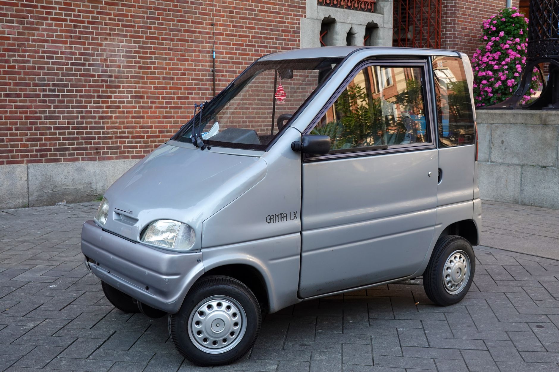 Silver microcar parked on a paved city street against a brick wall in daylight.