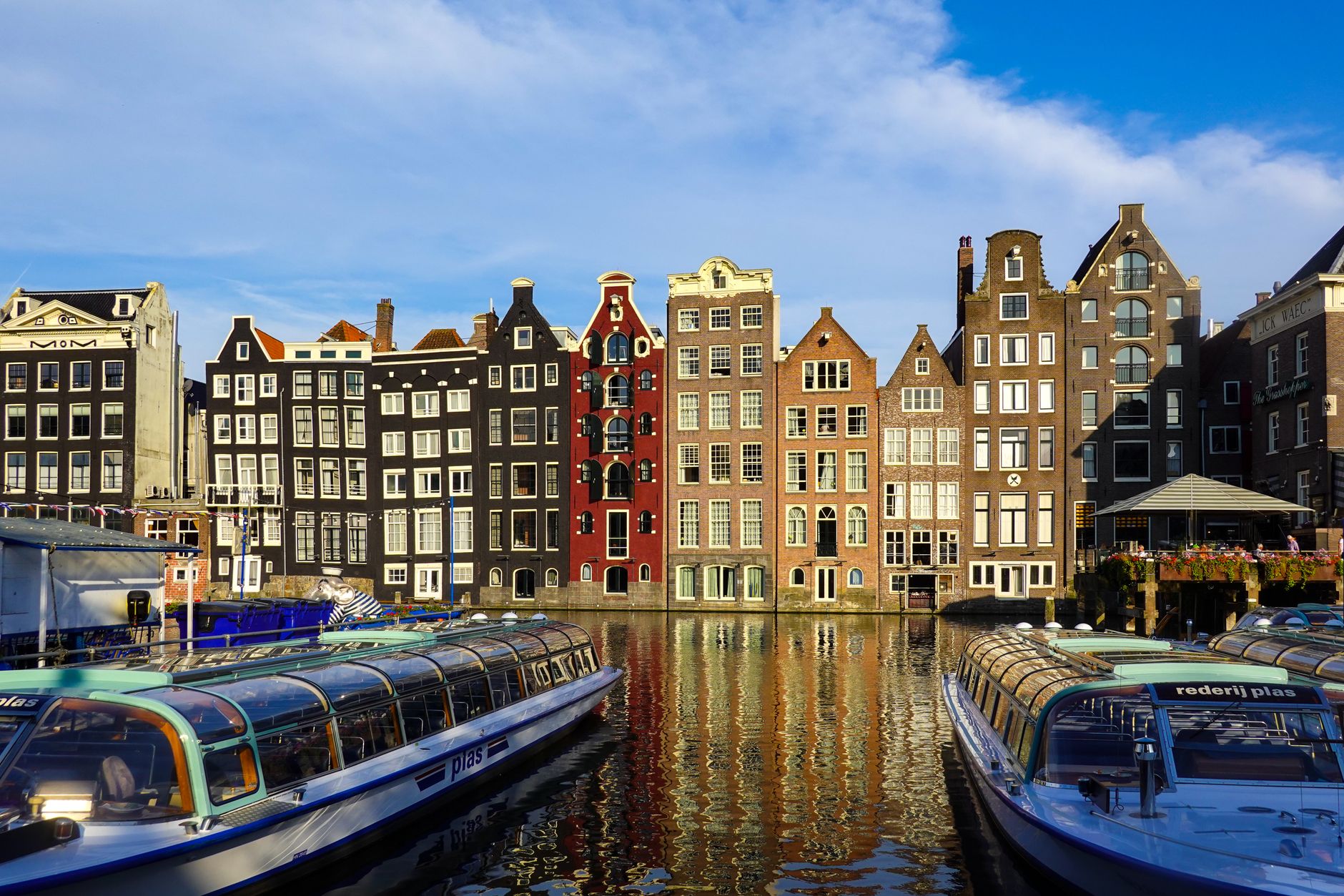 Colorful historic buildings along Amsterdam canals with boats in the foreground, reflecting in calm water.