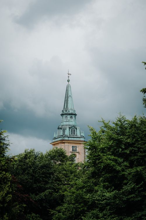 Free A picturesque view of a church tower emerging above verdant trees under a cloudy sky. Stock Photo