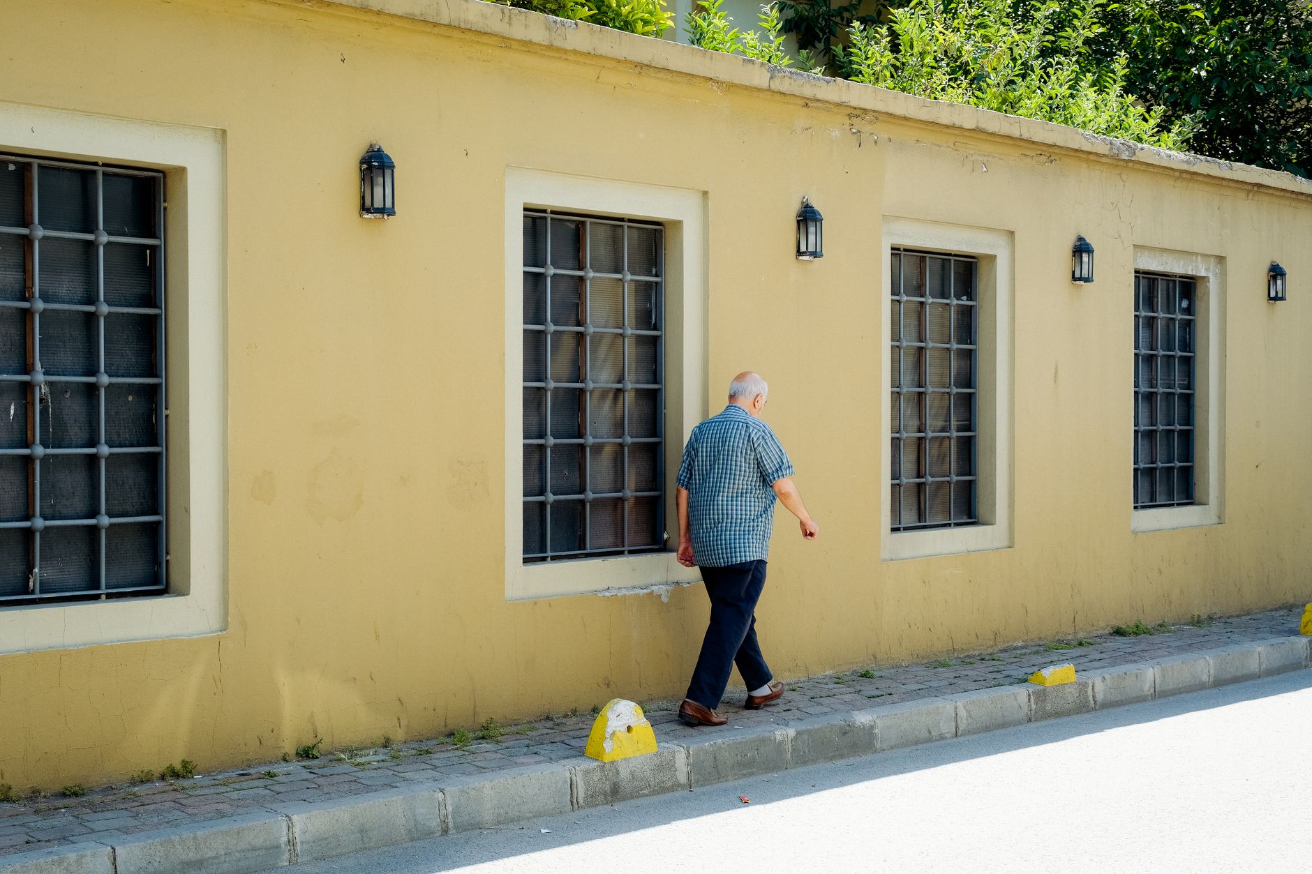 parkinson’s natural support — An elderly man walks on a sunny day along a yellow wall with high windows and lamps.