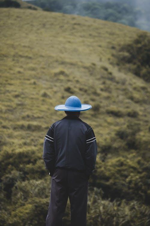 Free Back View of a Person Wearing Blue Hat while Standing in Front of the Green Mountain Stock Photo