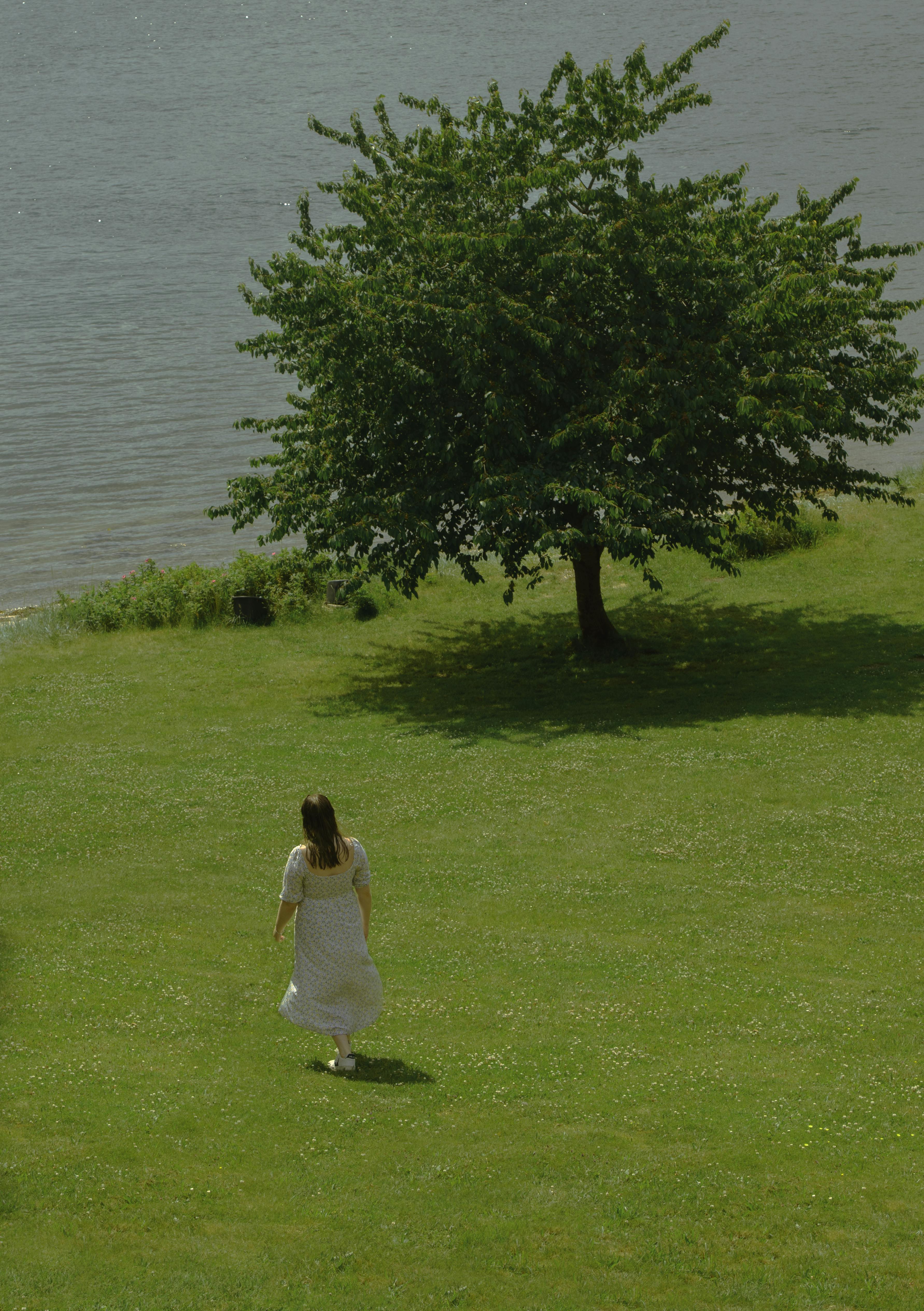 A woman in a flowing white dress walks across a lush green field, by a serene river.
