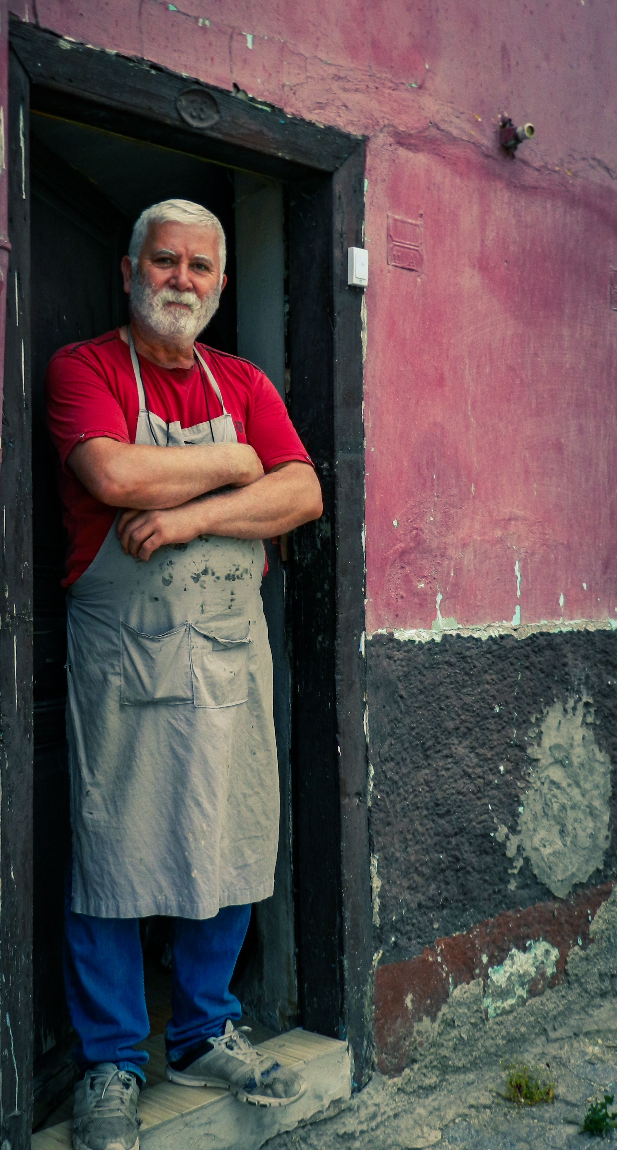 Elderly man with a beard and apron stands confidently in a doorway.