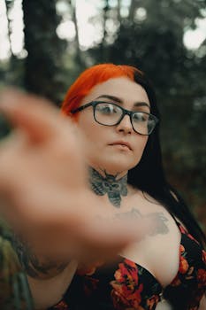 Close-up portrait of a tattooed woman with glasses in a natural setting, showcasing unique tattoos and red hair.