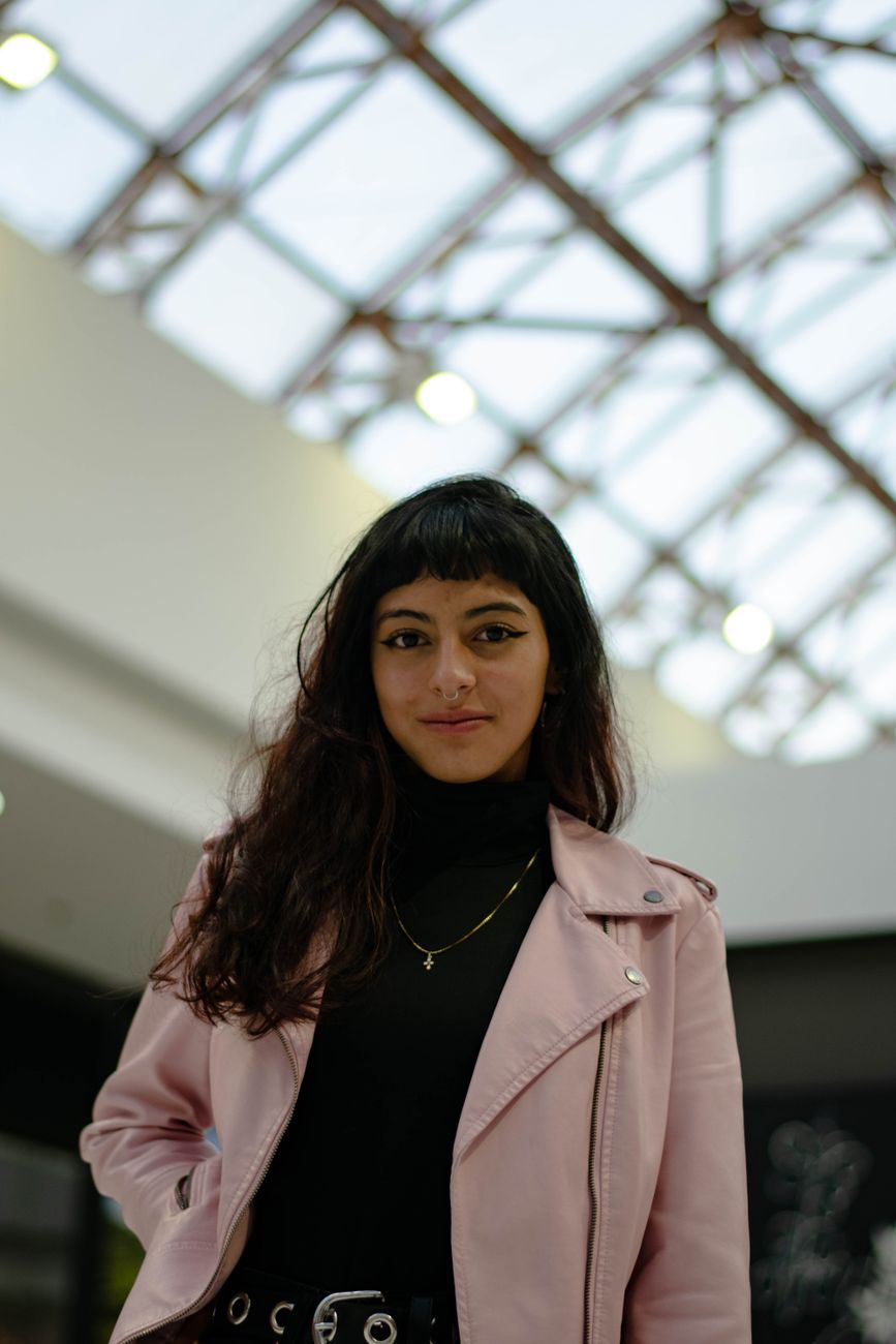 A fashionable woman poses confidently indoors wearing a pink jacket.