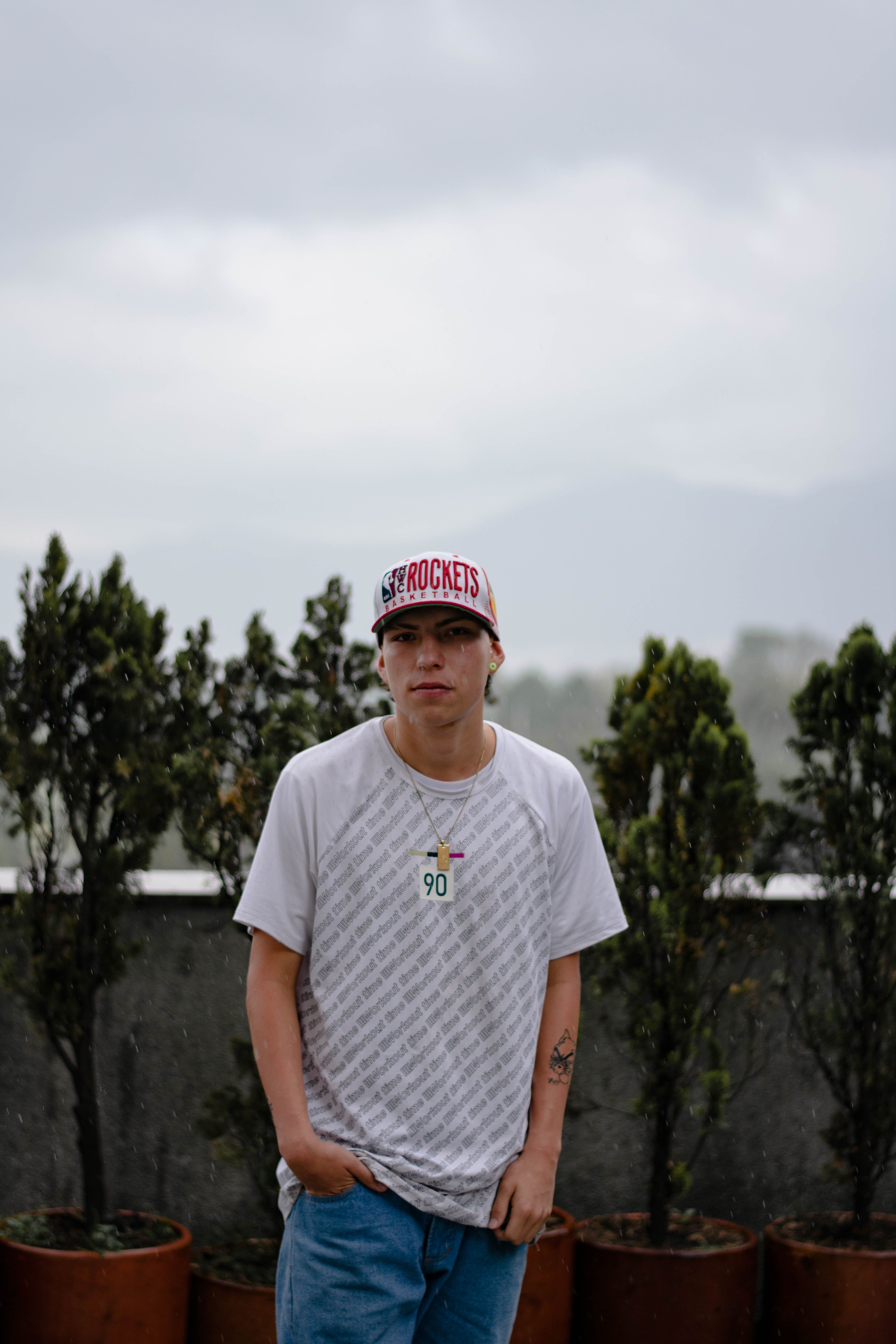 Young man in casual wear standing outdoors with lush greenery backdrop.