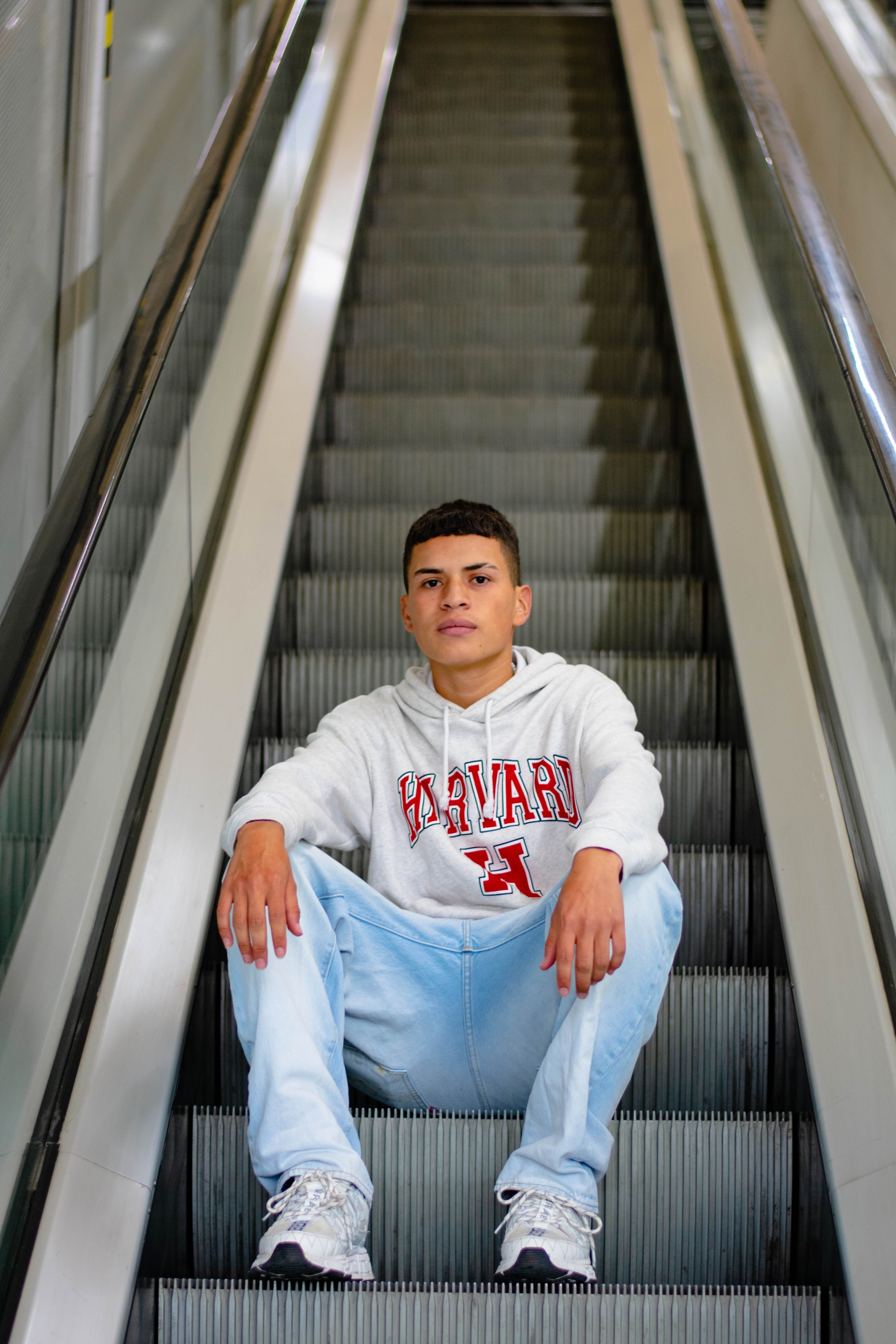 Teenage boy sitting on an escalator wearing a Harvard hoodie and jeans.