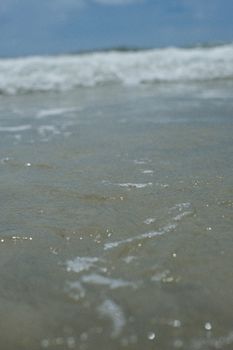 A serene close-up of gentle ocean waves and foam on a sandy beach under a clear blue sky.