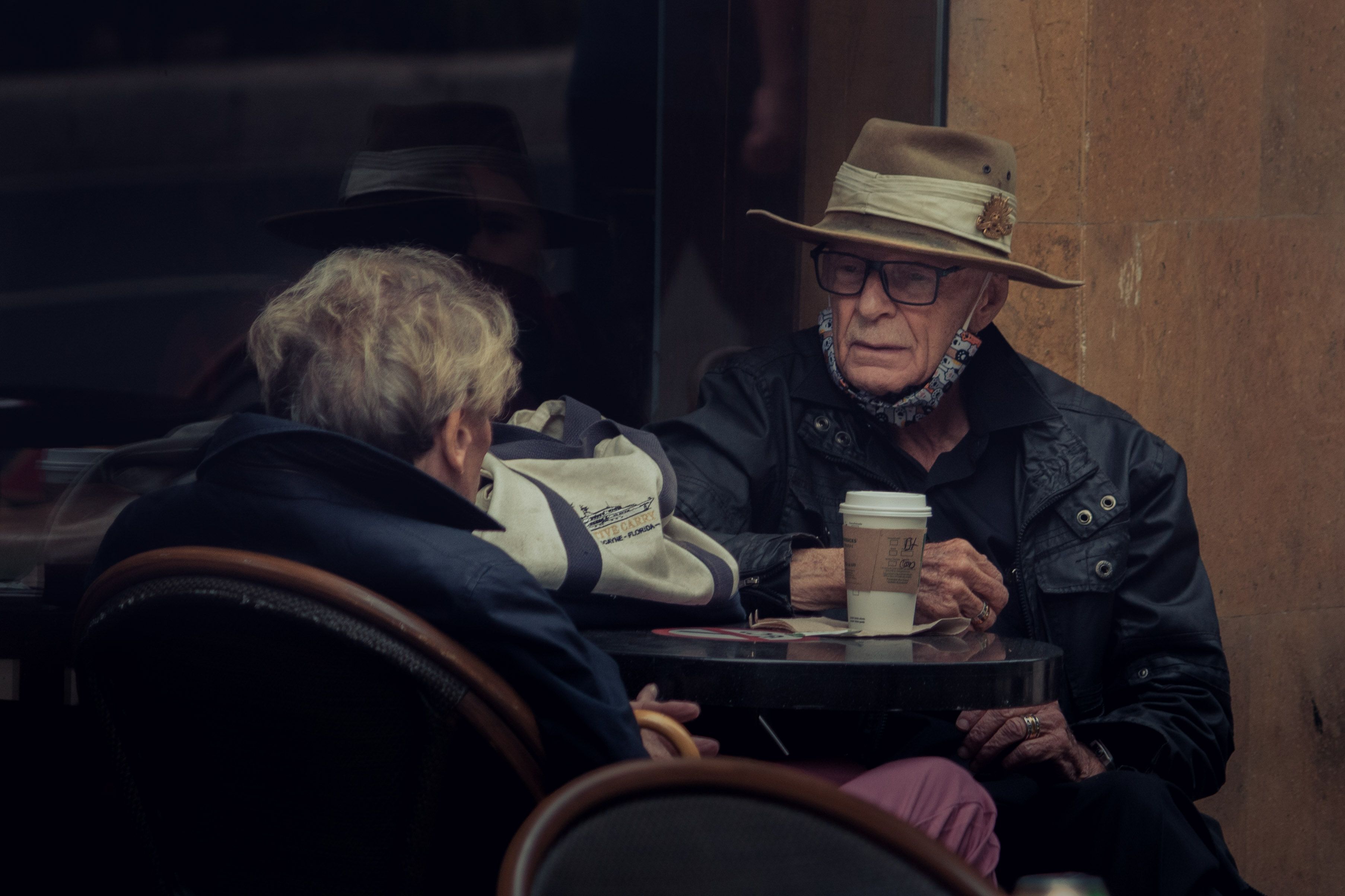 Elderly couple at an outdoor cafe sharing a coffee, wearing hats and jackets.