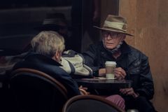 Elderly couple at an outdoor cafe sharing a coffee, wearing hats and jackets.