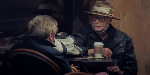 Elderly couple at an outdoor cafe sharing a coffee, wearing hats and jackets.