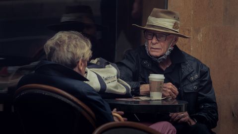 Elderly couple at an outdoor cafe sharing a coffee, wearing hats and jackets.