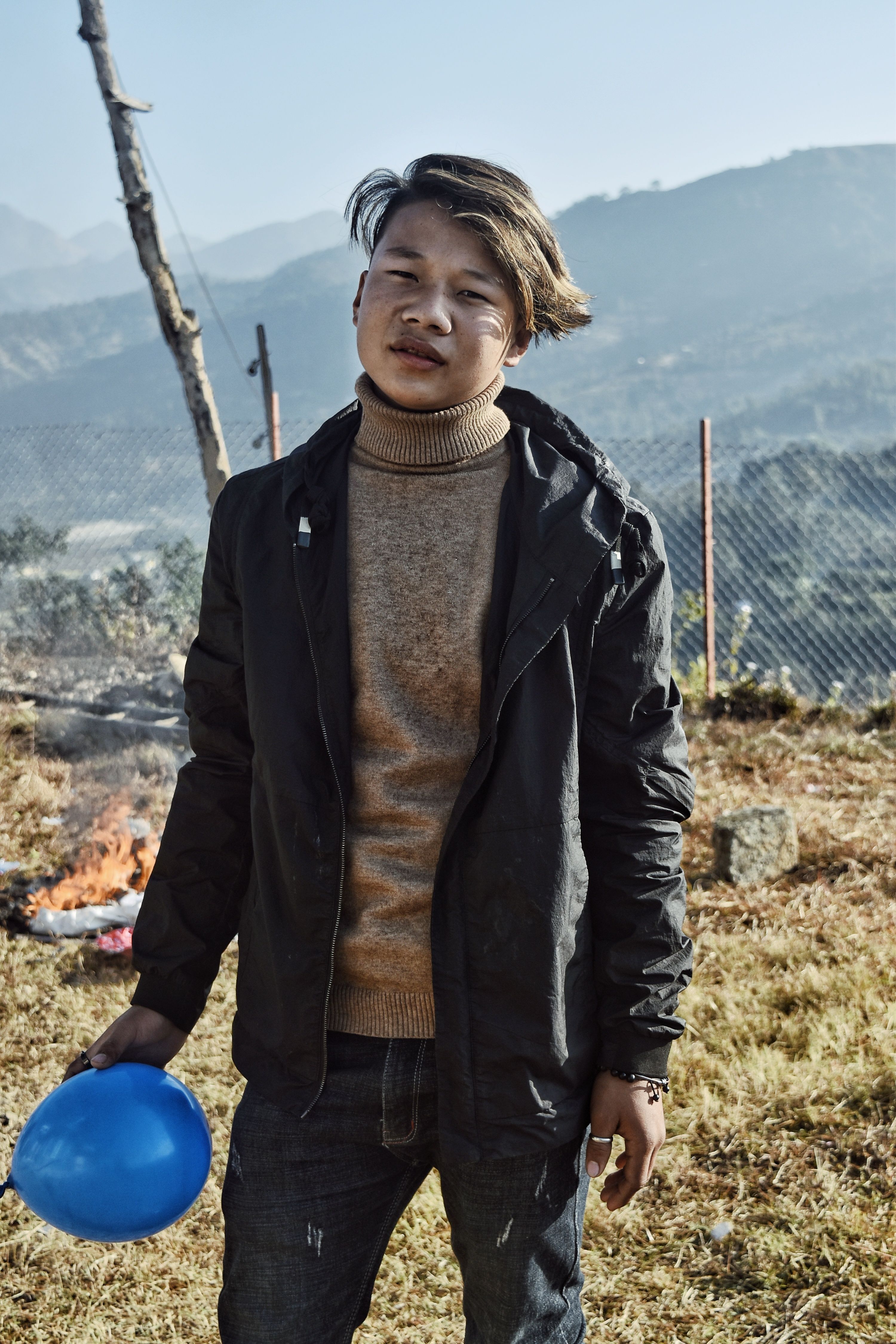 A young man in a black jacket holds a blue balloon outdoors on a sunny day.