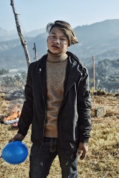 A young man in a black jacket holds a blue balloon outdoors on a sunny day.