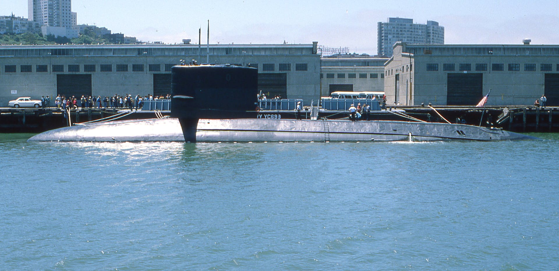 A naval submarine docked at an urban harbor with people on shore.