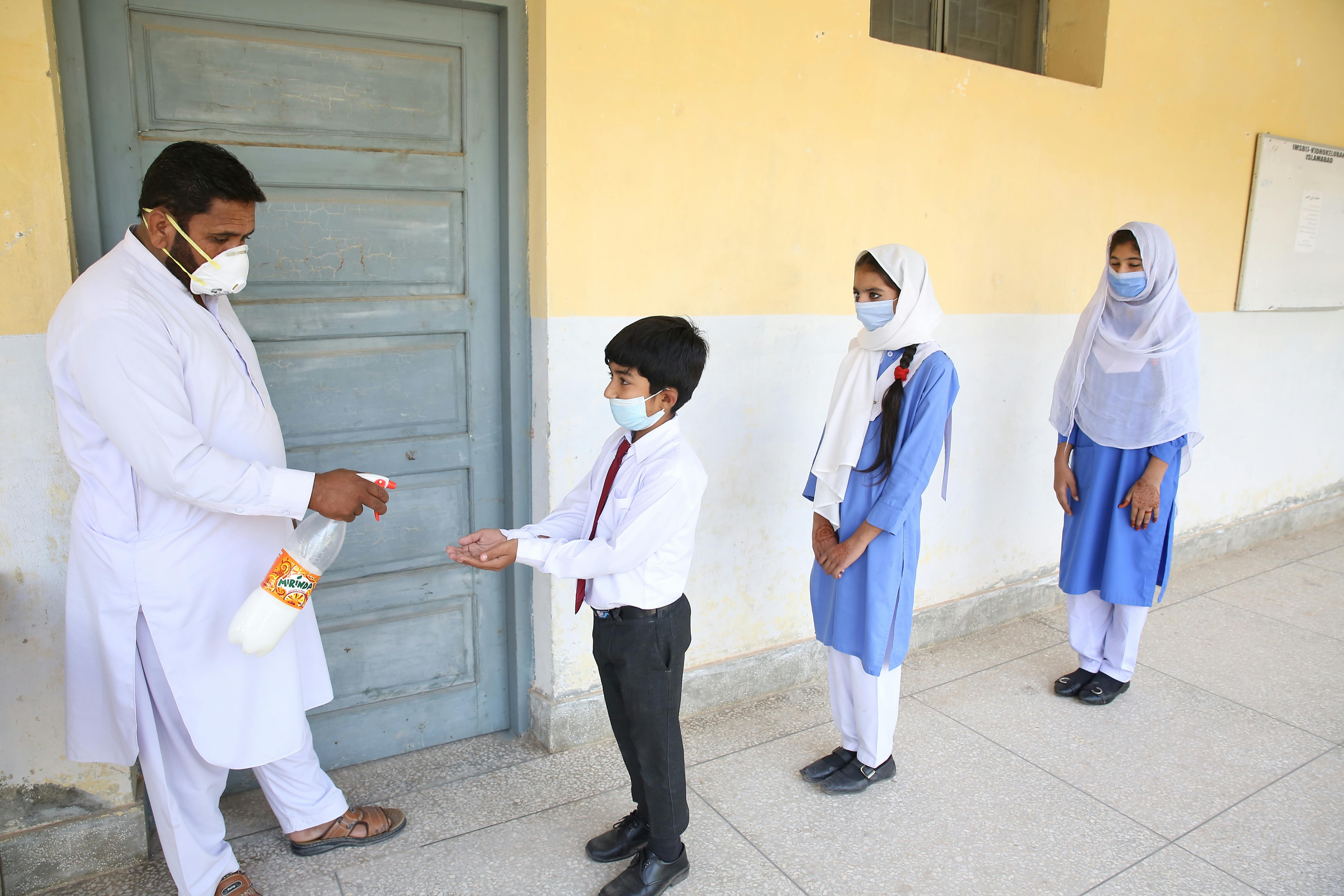 Students wearing masks use sanitizer at a school entrance, ensuring hygiene and safety.