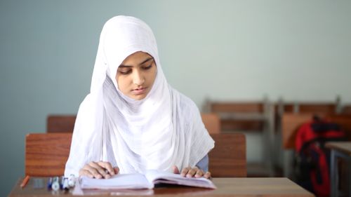 Free Young girl in hijab reading at a desk in a classroom. Focused and studious. Stock Photo