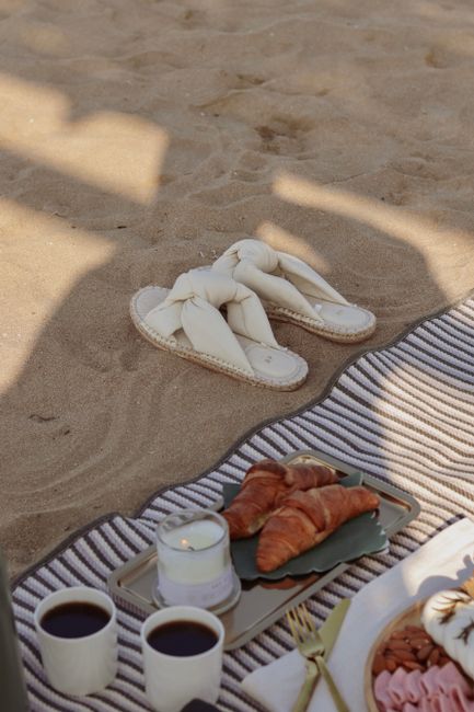 A cozy beach picnic setting with croissants, coffee, and a candle on sand.
