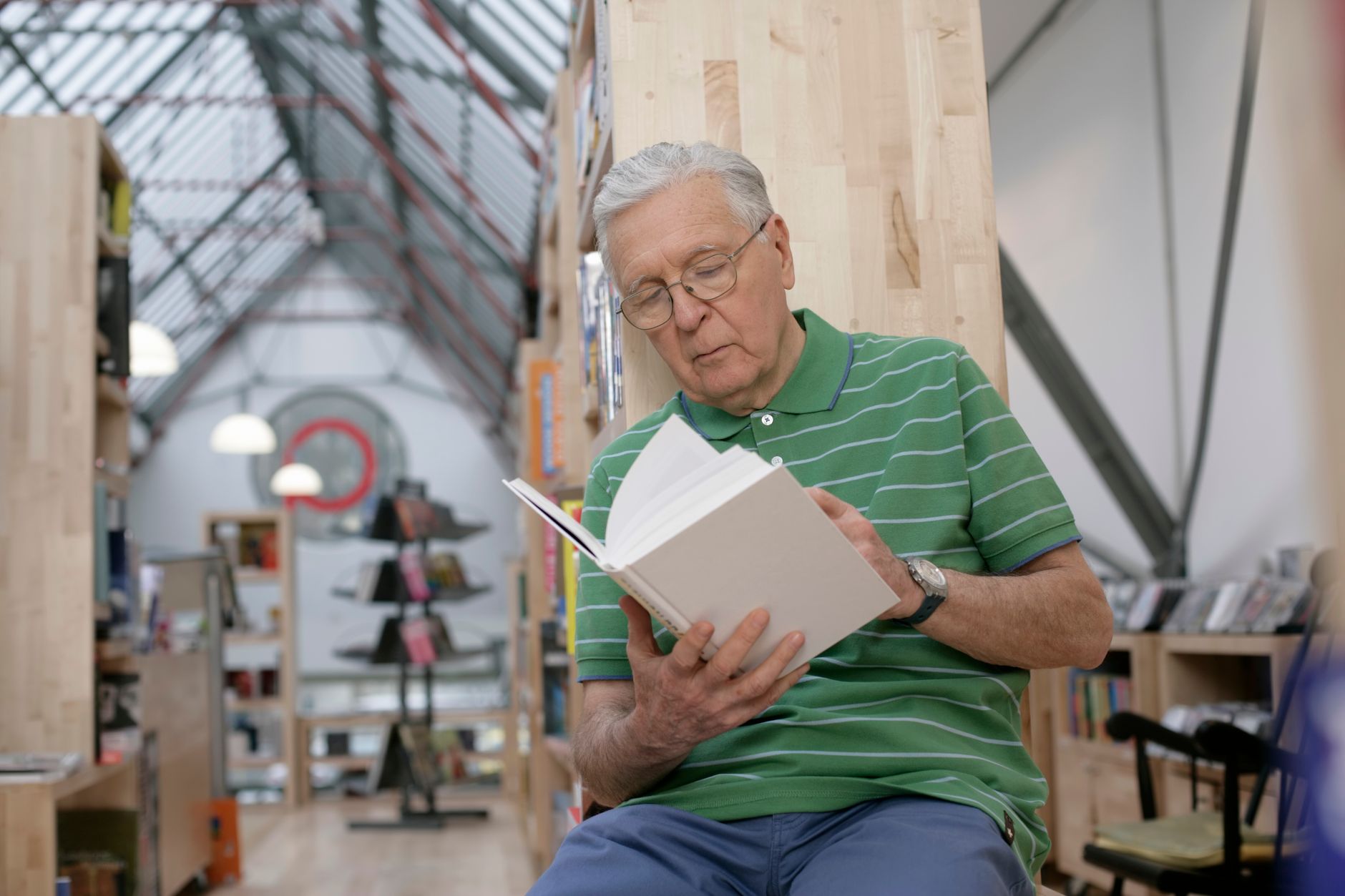 Elderly man reading a book in a bright, modern library setting, wearing glasses and focused.