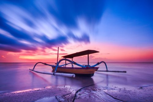 Free A traditional fishing boat on a tranquil seashore during a vibrant sunrise with dramatic clouds. Stock Photo