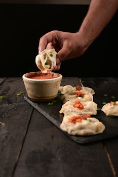 A close-up of hand dipping dumpling into sauce on rustic table with dumplings presented.