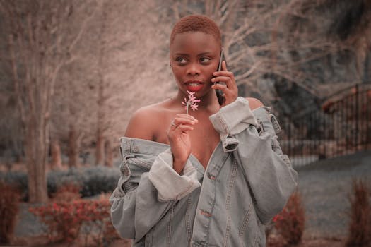 Portrait of a stylish woman in a denim jacket talking on the phone while holding a flower outdoors.
