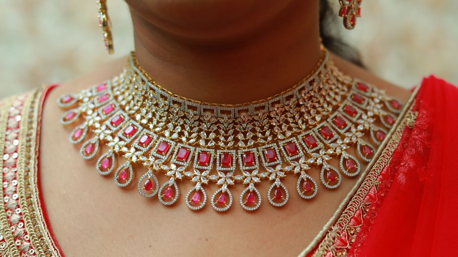 A red ruby pendant necklace and matching ruby stud earrings in gold arranged on a neutral editorial background