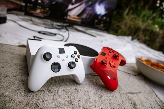 White and red wireless gaming controllers on a textured surface near chips and a TV screen.