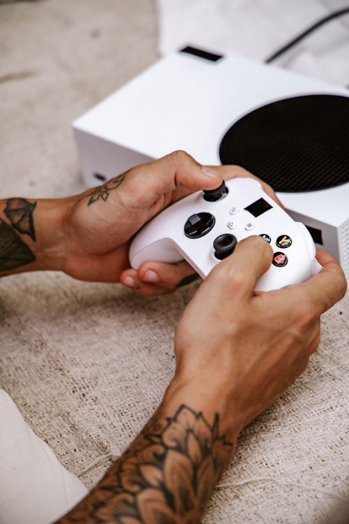 Free A close-up of tattooed hands gripping a white gaming controller with a console in the background on a textured surface. Stock Photo