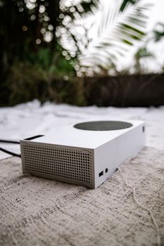 A sleek white game console placed on a textured outdoor rug, surrounded by nature.