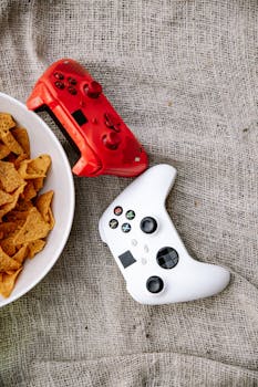 Close-up of red and white game controllers next to a bowl of chips on textured fabric.