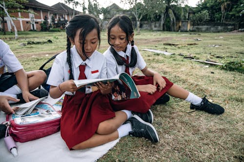Two Female Students Sitting on the Field while Reading Books