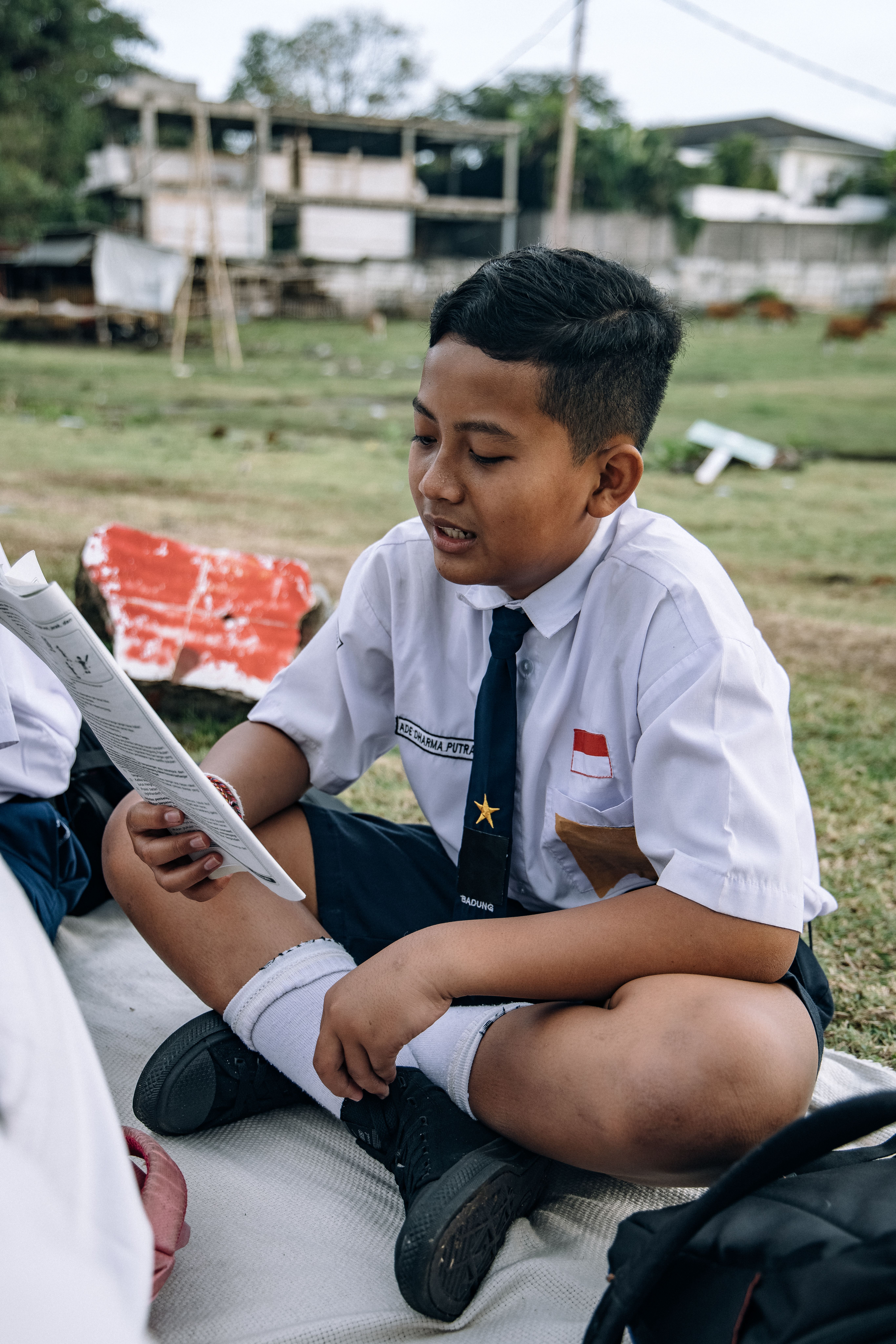 Seorang siswa berseragam sedang membaca buku di luar ruangan.
