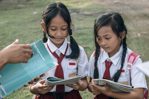 Two young girls in school uniforms reading outdoors with a book in hand, captured in natural daylight.
