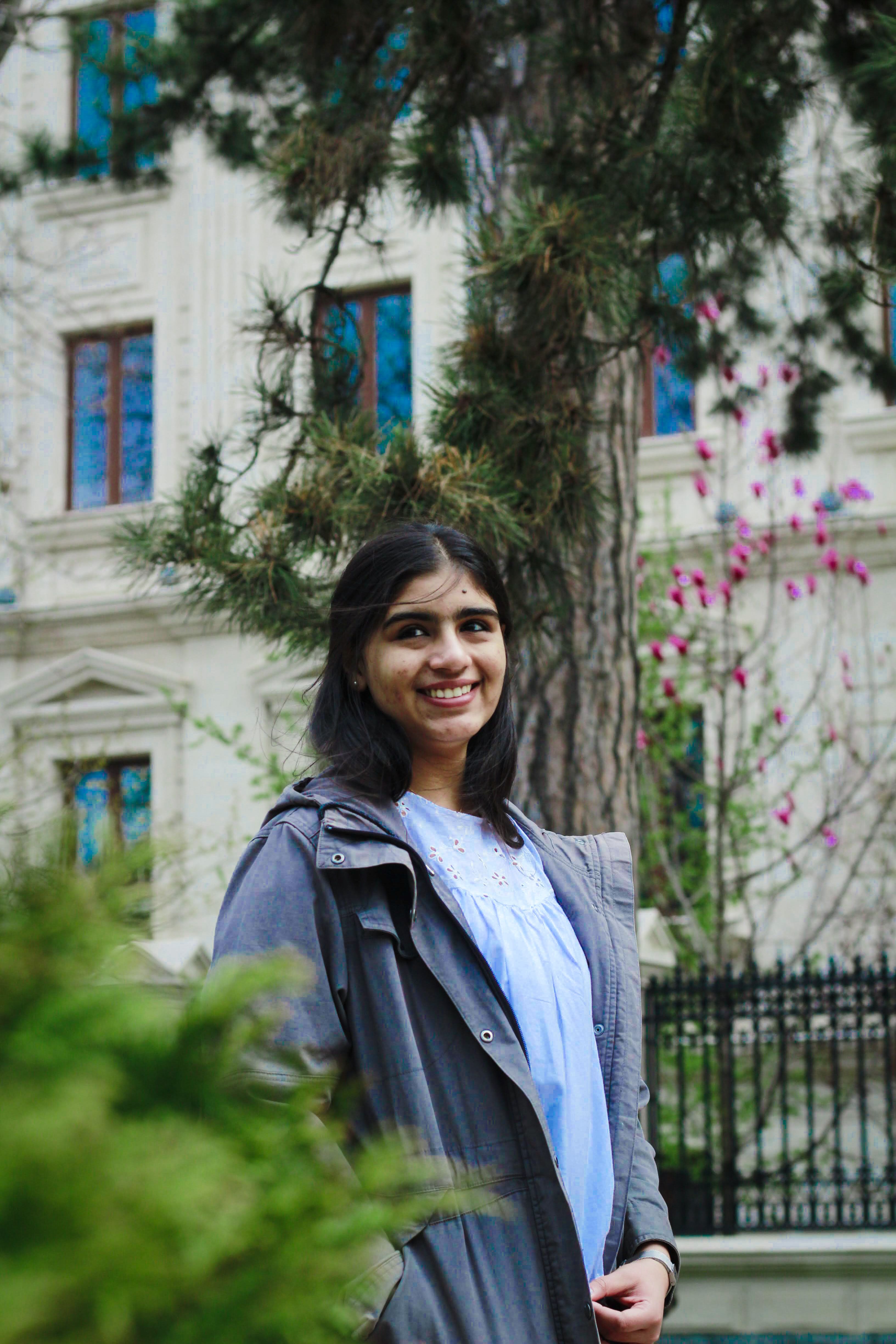 A smiling woman in a blue jacket stands outdoors with blooming flowers in the backdrop.