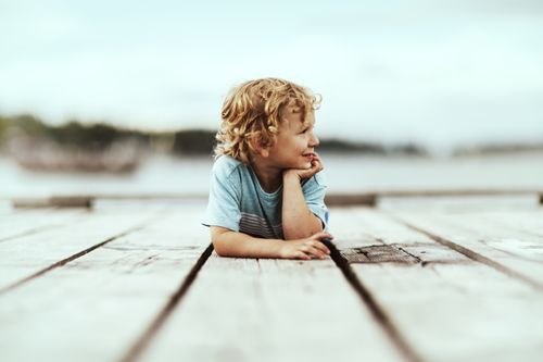 Free A young boy with curly hair lying on a wooden pier, enjoying a serene summer day. Stock Photo