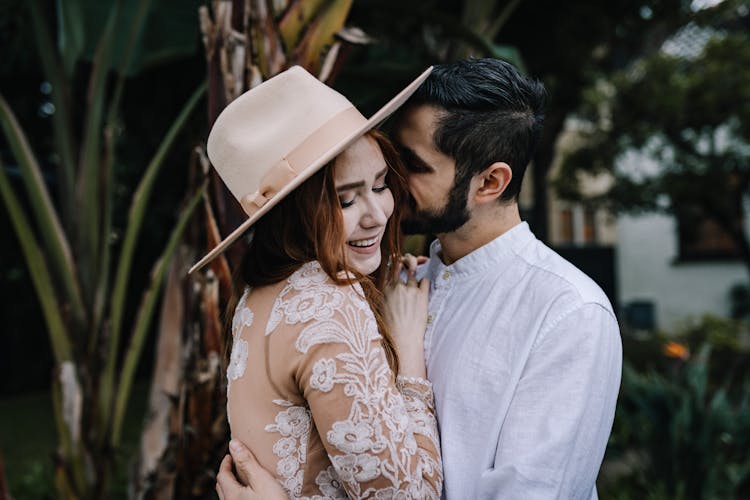 Young Woman In A Brown Embroidered Floral Dress And Hat Being Kissed By A Man Embracing Her