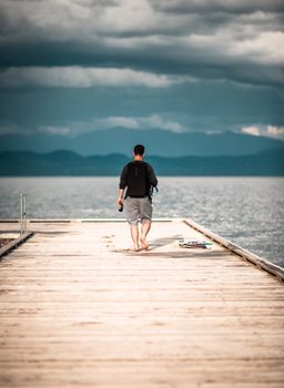 A man walks barefoot on a wooden dock leading to a calm sea under a cloudy sky.